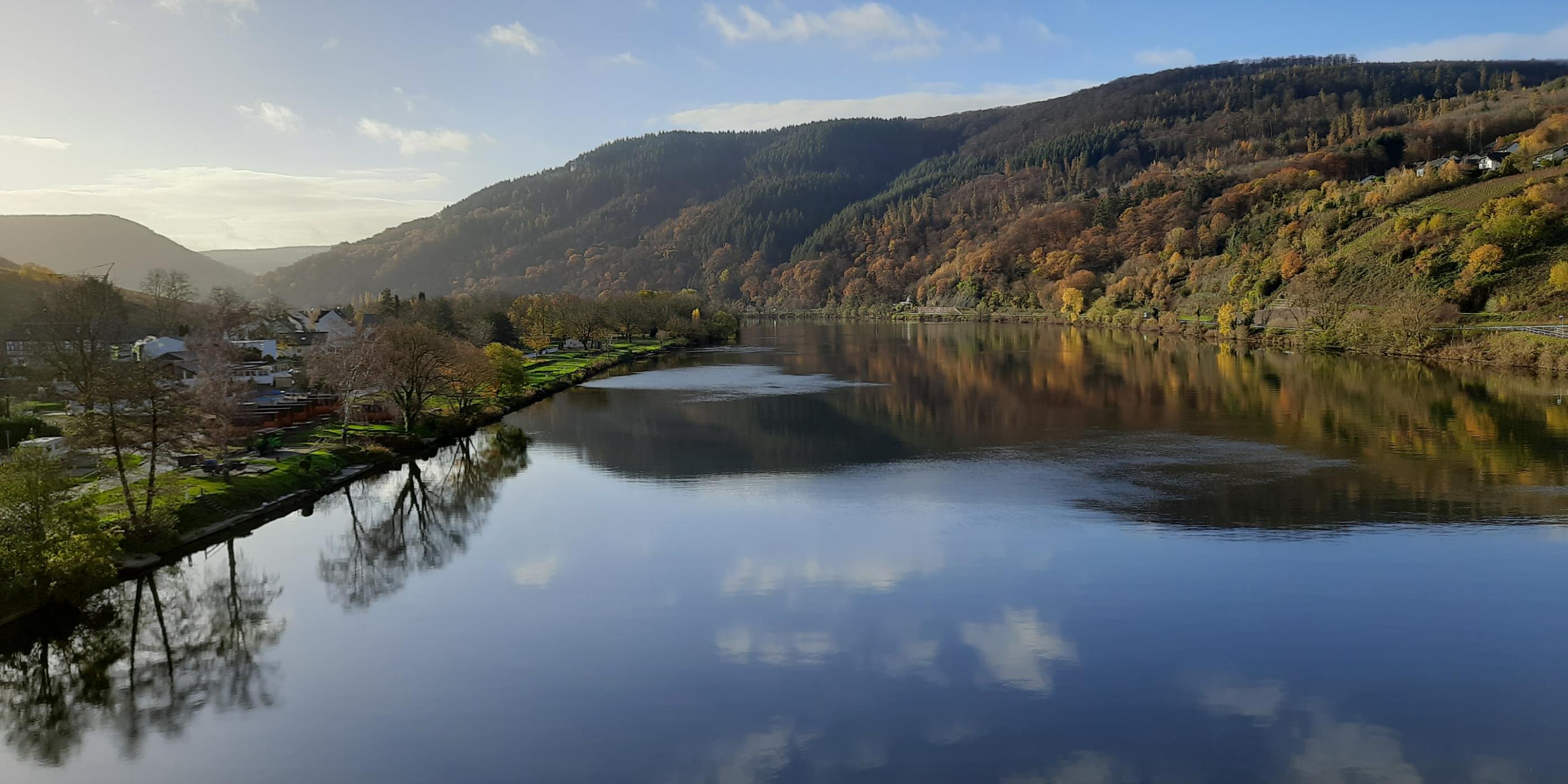 Blick auf die Mosel. Rechts und links sieht man Landschaft. Der Himmel ist blau mit ein paar Wölkchen, die sich im Wasser spiegeln